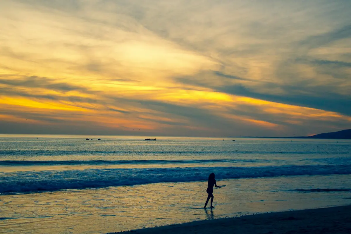 Silhouetted person walking along a sandy beach at sunset with calm ocean waves and a colorful sky.