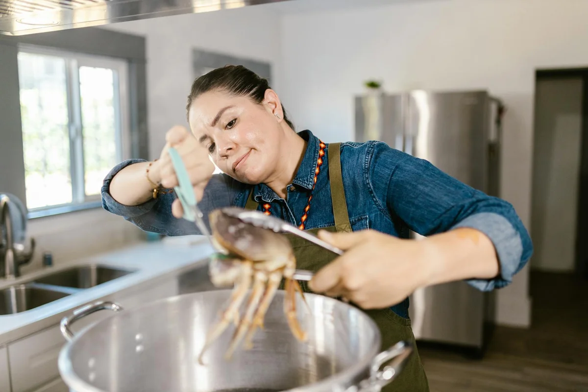Person in a denim shirt and apron carefully handling a hermit crab with tongs over a large stainless steel pot in a kitchen