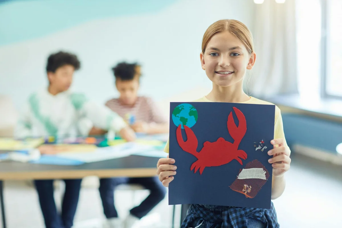 A smiling girl in a bright classroom holds up a blue poster depicting a red hermit crab and a globe, with two other students drawing in the blurred background.