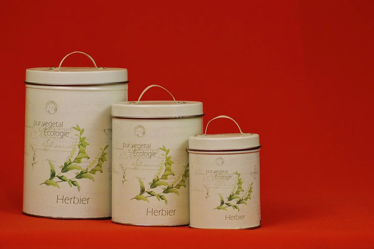 Three white metal storage tins with lids and handles, arranged on a red background, featuring leafy plant illustrations.