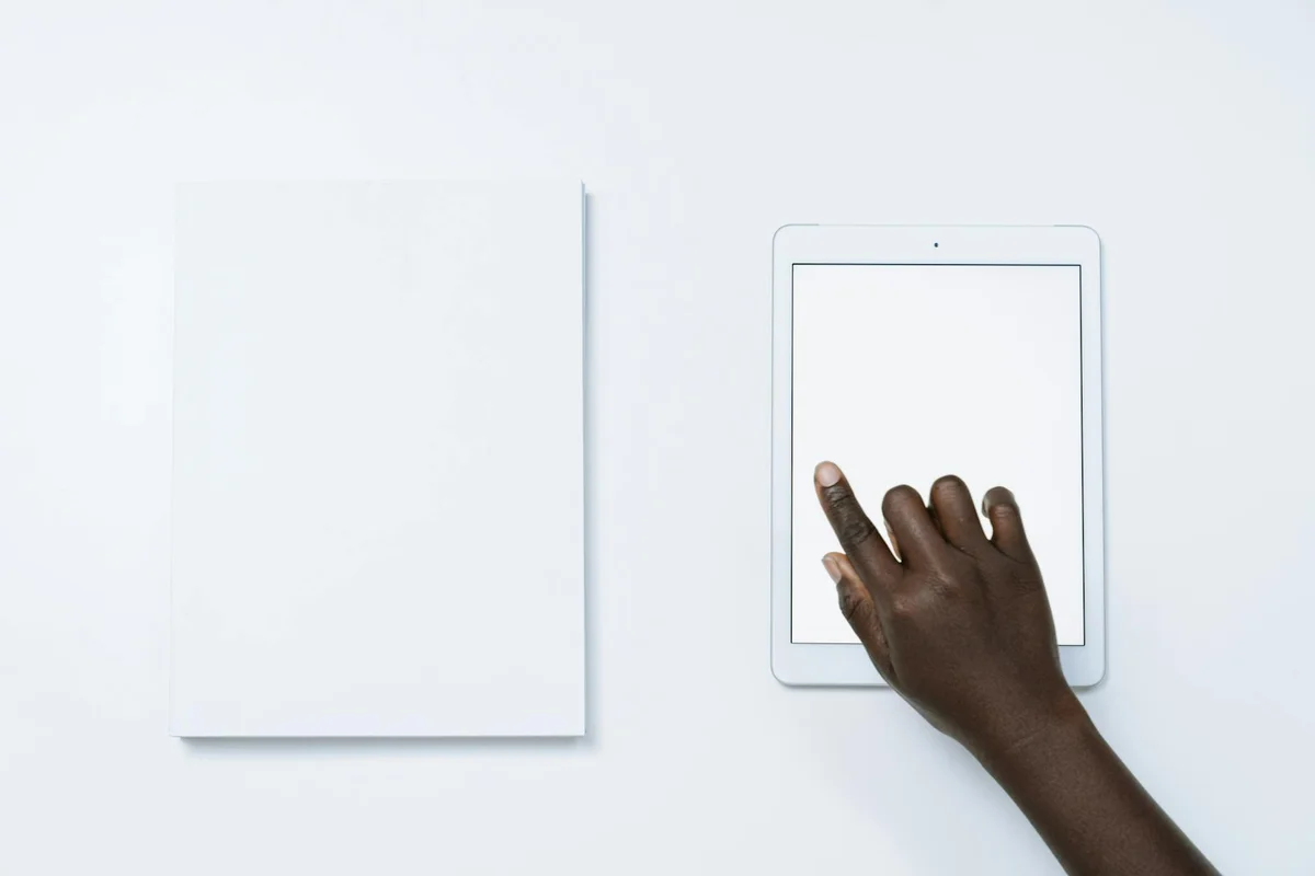 Dark-skinned hand using a tablet on a white desk next to a blank notebook.