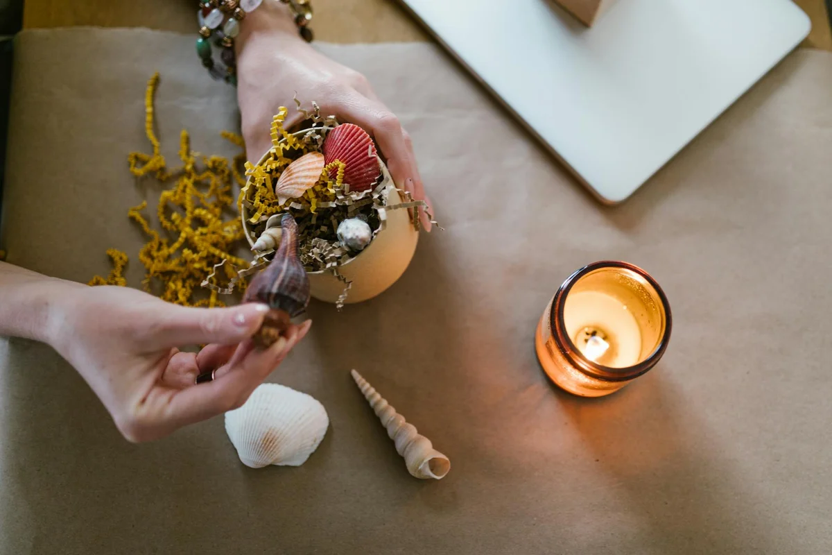 Close-up of hands arranging shells and decorative items on a tabletop for hermit crab care, with a small candle burning nearby.