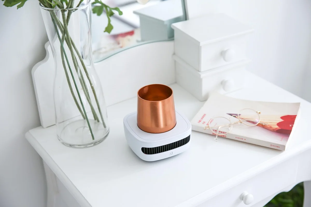 Close-up of a compact white and copper heating device on a white dresser, with a plant and a book nearby, illustrating equipment used to regulate temperature.