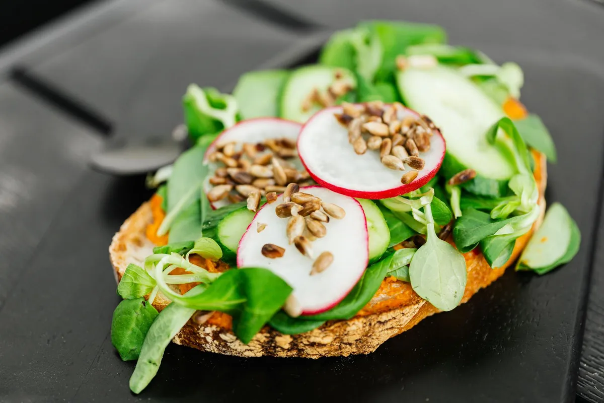 Open-faced toast topped with radish slices, cucumber rounds, and leafy greens, sprinkled with seeds, on a dark surface.