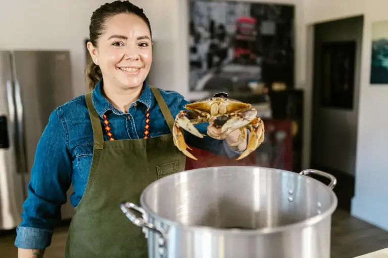 Smiling store employee wearing a denim shirt and green apron holds a hermit crab over a large stainless steel pot inside a pet shop.