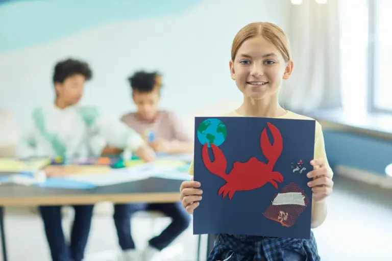 Young girl holding a blue poster featuring a red crab in a bright classroom, with other students working in the background.